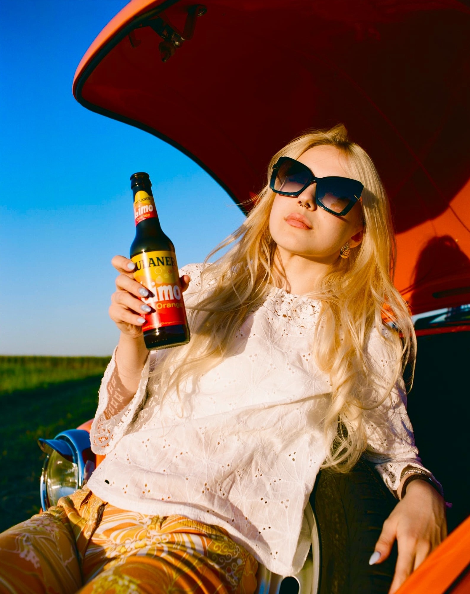 Maria in sunglasses leaning against a red vintage Beetle, bottle in hand