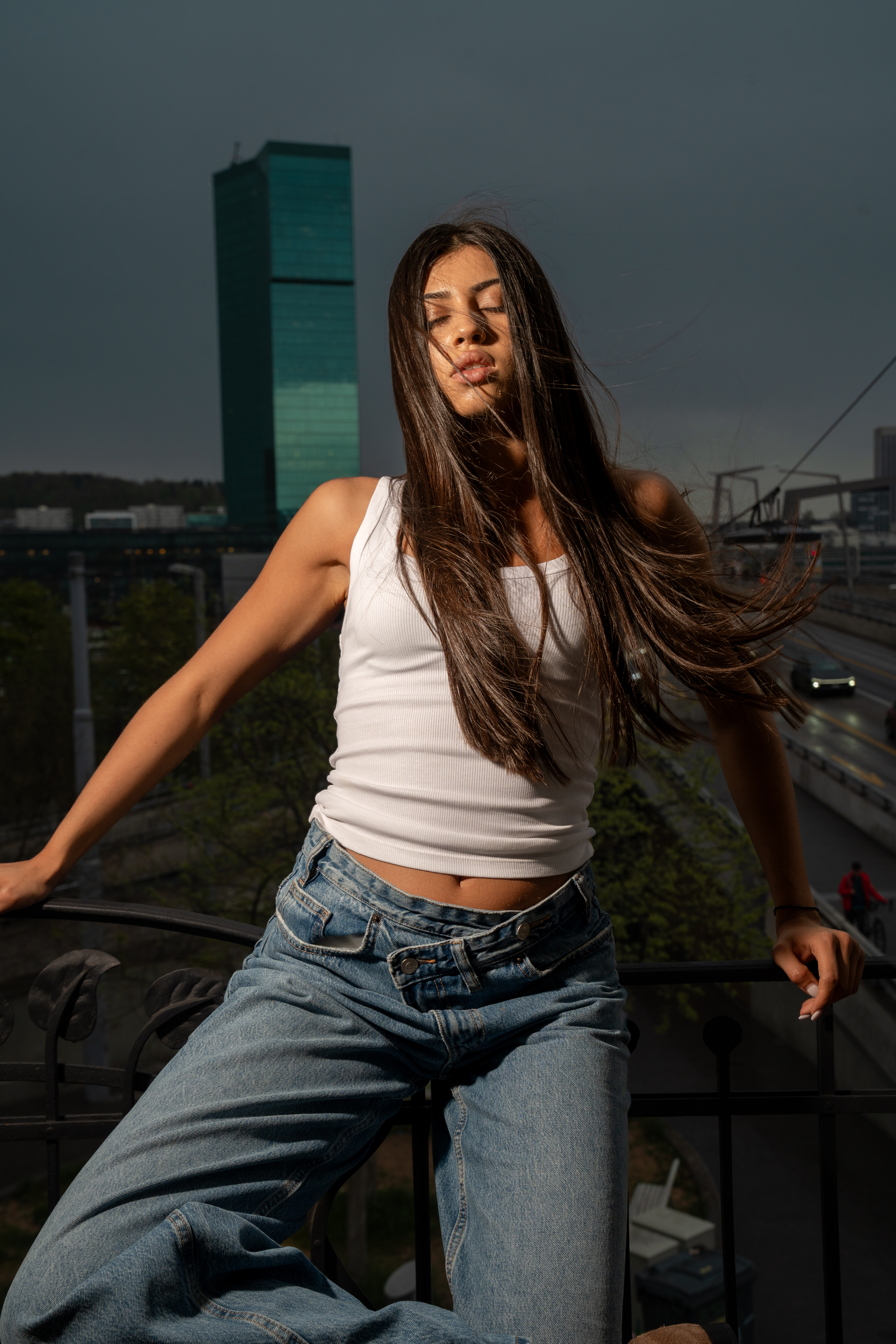 Model leaning on a balcony railing against a dark sky and a green glass tower